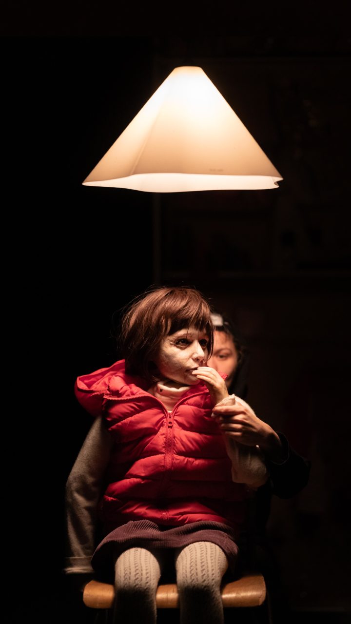 Une marionnette représentant une petite fille avec un gilet sans manches rouge. Au-dessus d'elle une lampe. Derrière elle, on devine la marionnettiste.