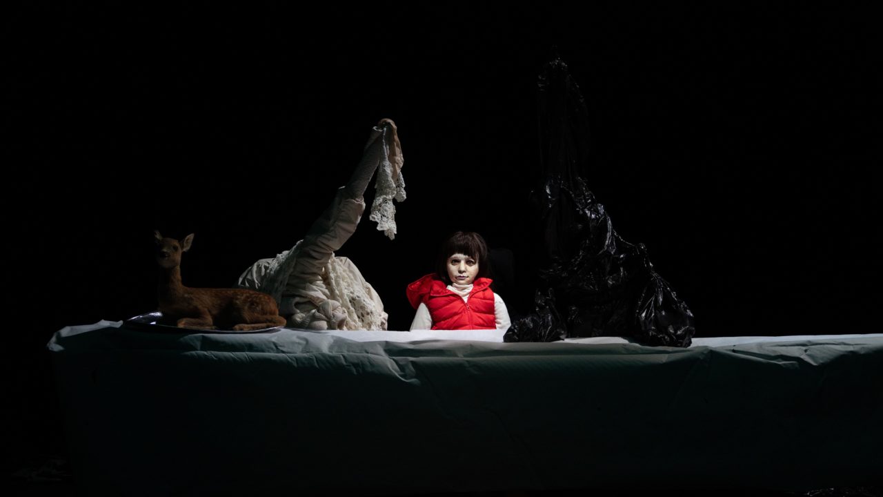 Une marionnette représentant une petite fille avec un gilet sans manches rouge. Elle se trouve à côté de grands objets noirs et blancs et d'un faon.