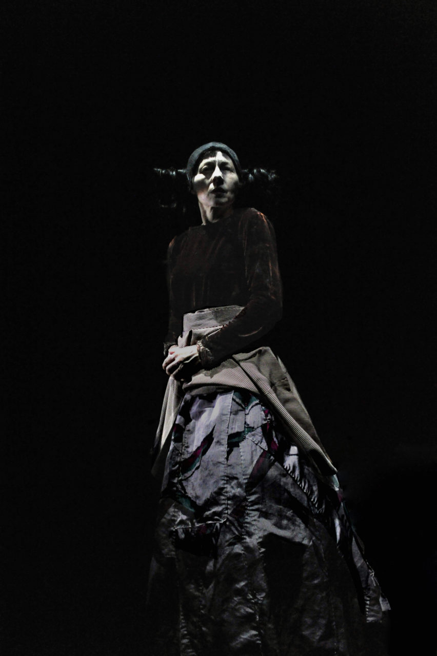 Un danseuse se tient debout avec un pull brun et une jupe patchwork. Elle regarde l'horizon.