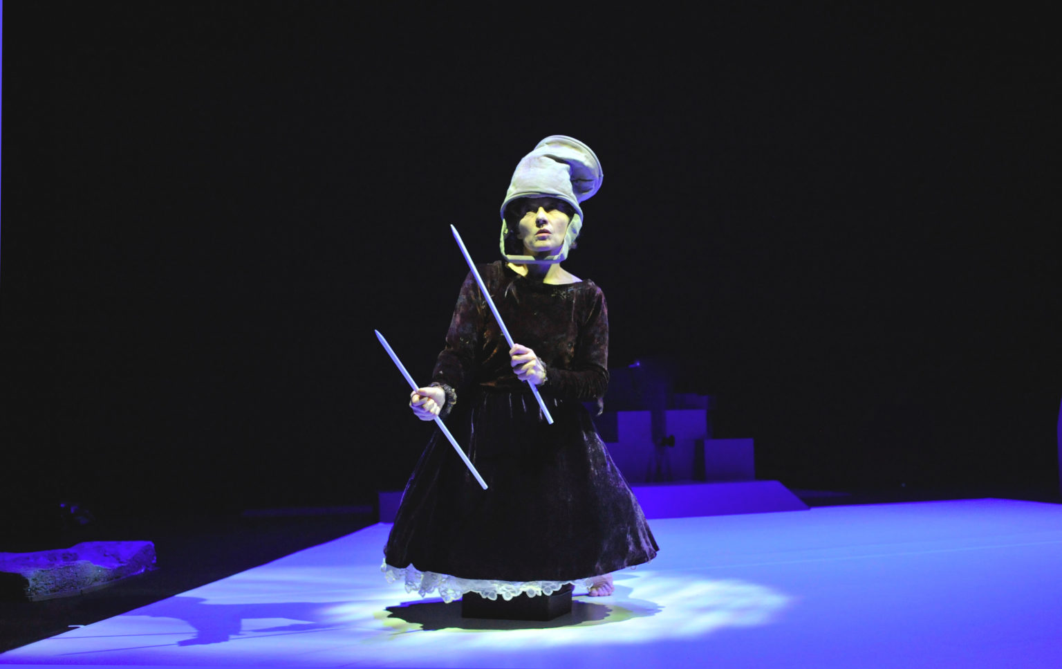 Une danseuse en robe marron est à genou sur scène. Ell porte un chapeau et elle à une baguette dans chaque main qu'elle tient devant elle.
