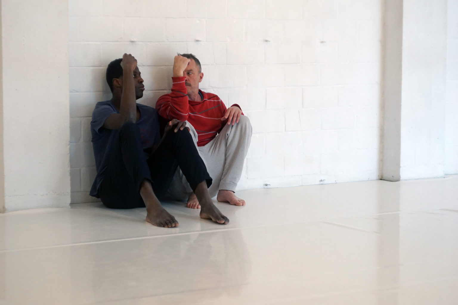 2 personnes sont assises par terre, dos contre un mure blanc, en train de discuter.