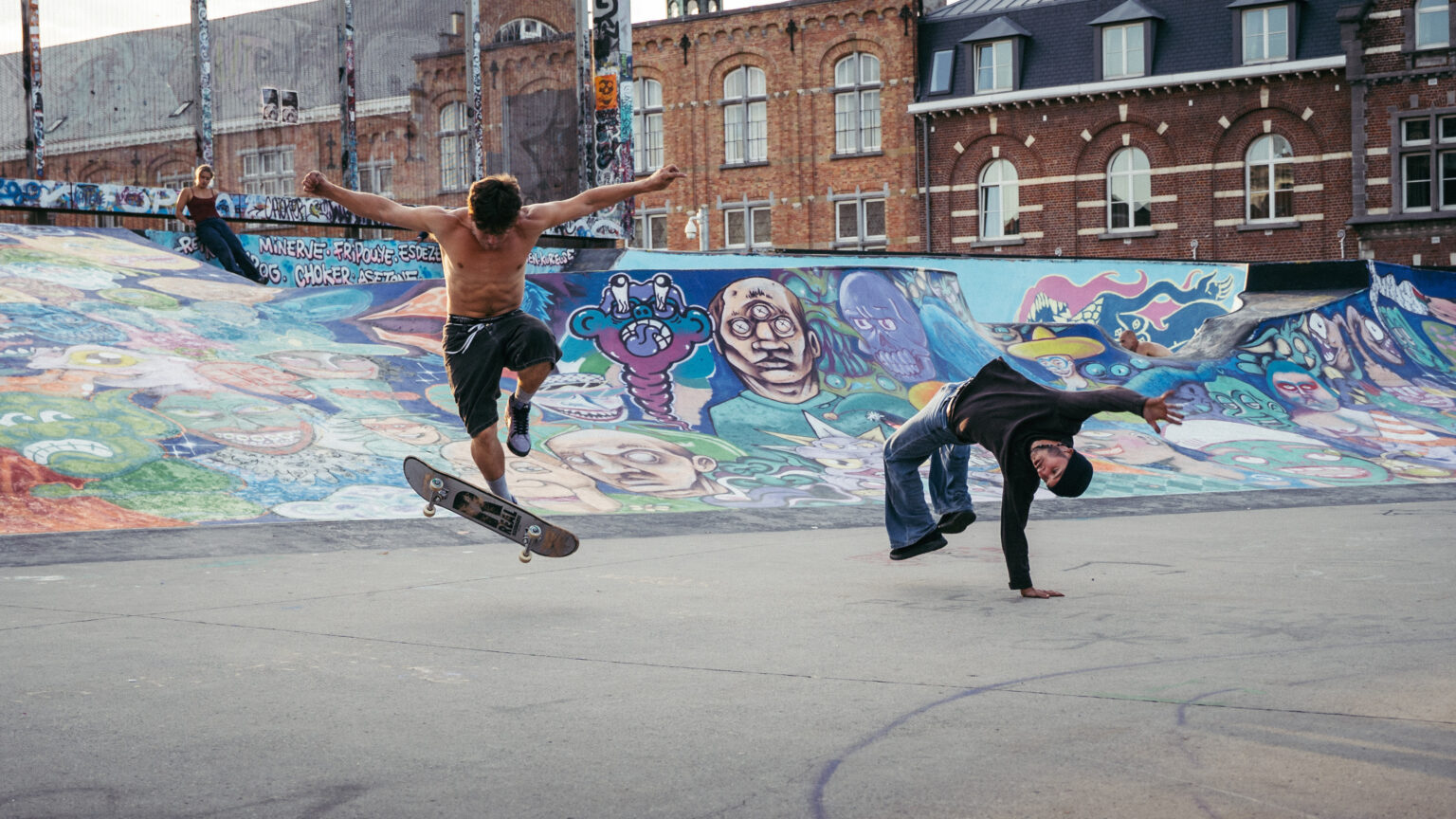 Un skateur fait un saut pendant qu'un danseur de breakdance fait une pirouette, une main sur le sol. La scène se passe au Skatepark des Ursulines.