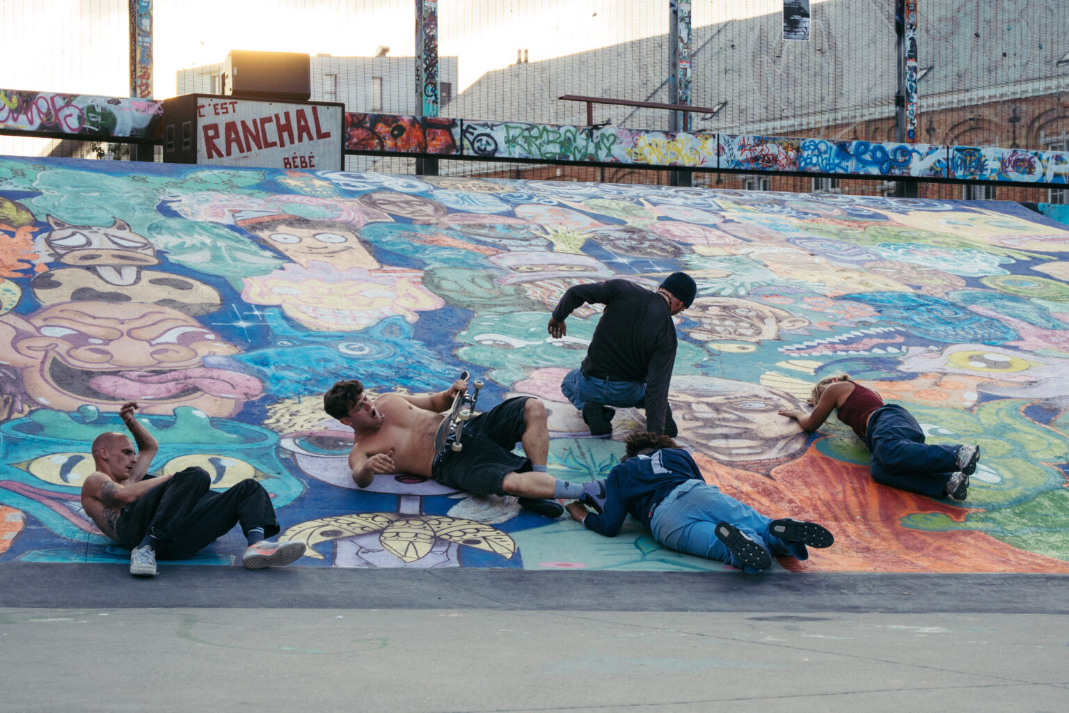5 danseurs et skateurs sont couchés sur le sol du skatepark, qui est taggué de fresques colorées. Le soleil décline au loin. La scène se passe au Skatepark des Ursulines.