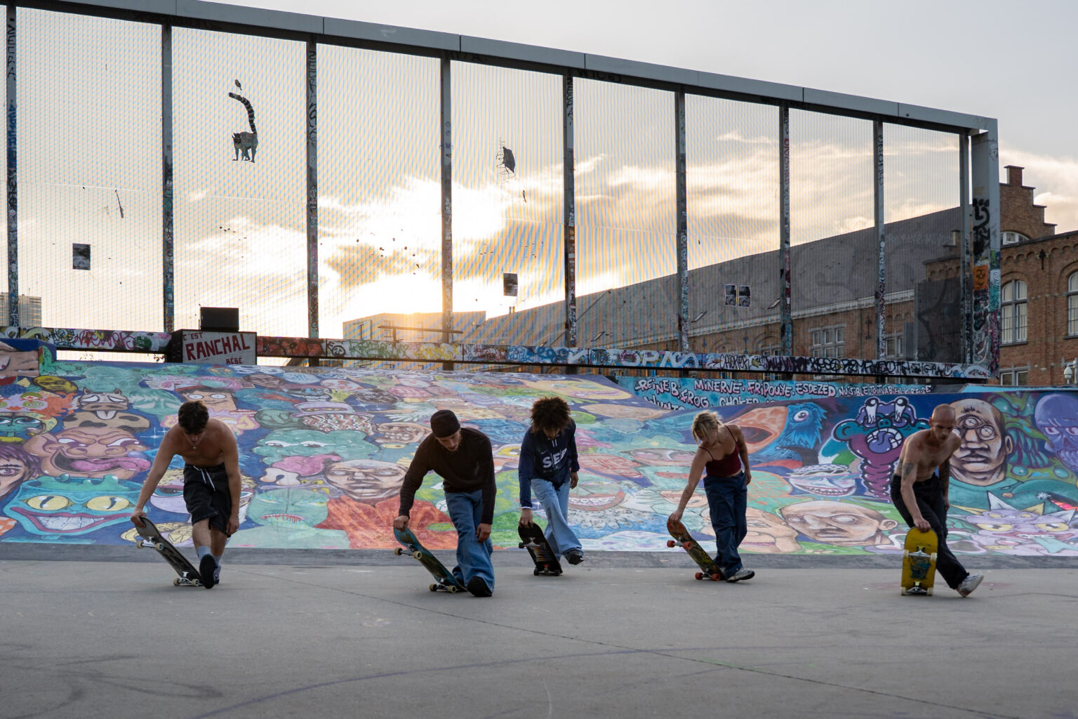 5 danseurs et skateurs sont penchés et tiennent un skateboard en mains. Le soleil décline au loin. La scène se passe au Skatepark des Ursulines.