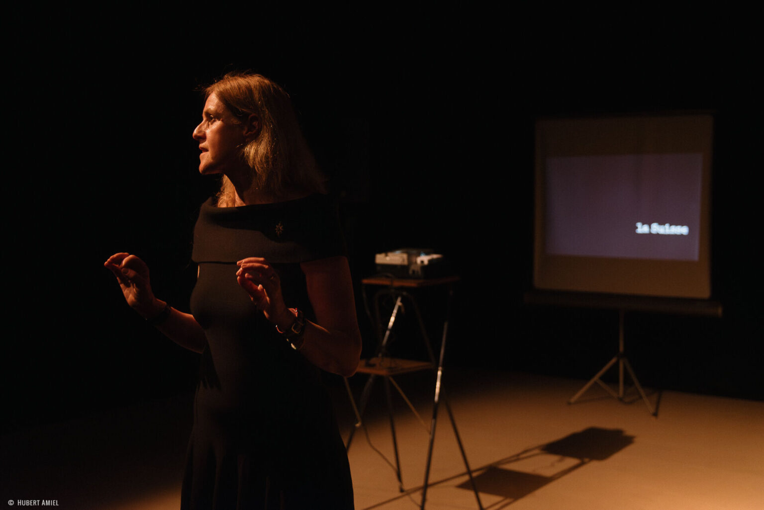 Geneviève Damas se tient debout. Derrière elle, se trouve un écran noir affichant "