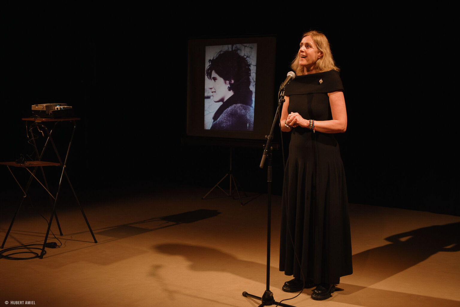 Geneviève Damas se tient debout, les mains jointes, derrière un micro. A coté d'elle, se trouve un écran montrant la photo d'une jeune femme.