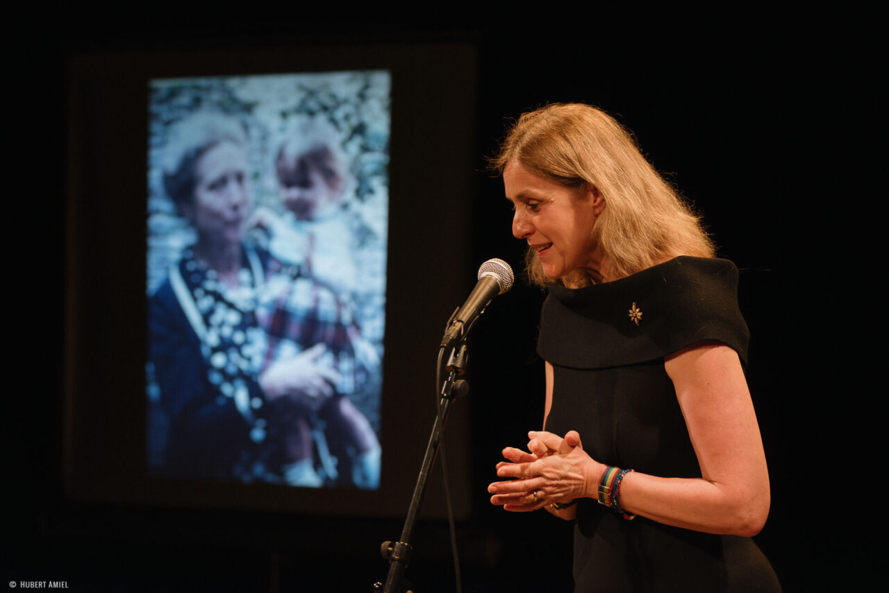 Geneviève Damas se tient debout, les mains jointes, derrière un micro. A coté d'elle, se trouve un écran montrant la photo d'une femme portant une petite fille.