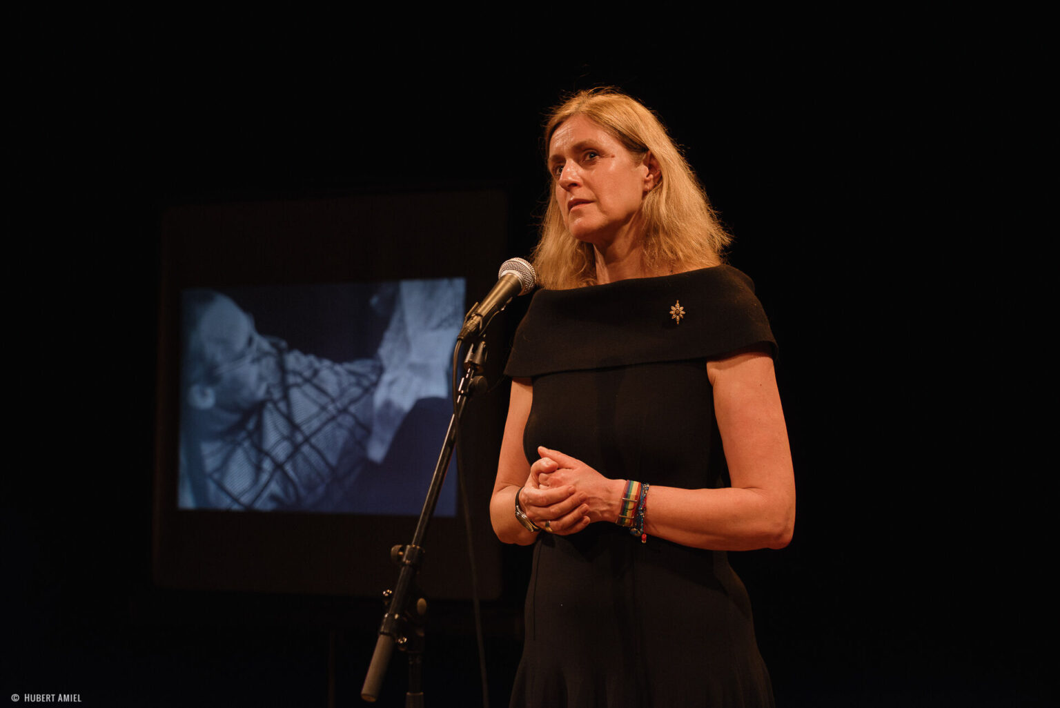 Geneviève Damas se tient debout, les mains jointes, derrière un micro. A coté d'elle, se trouve un écran montrant la photo d'un homme assis.