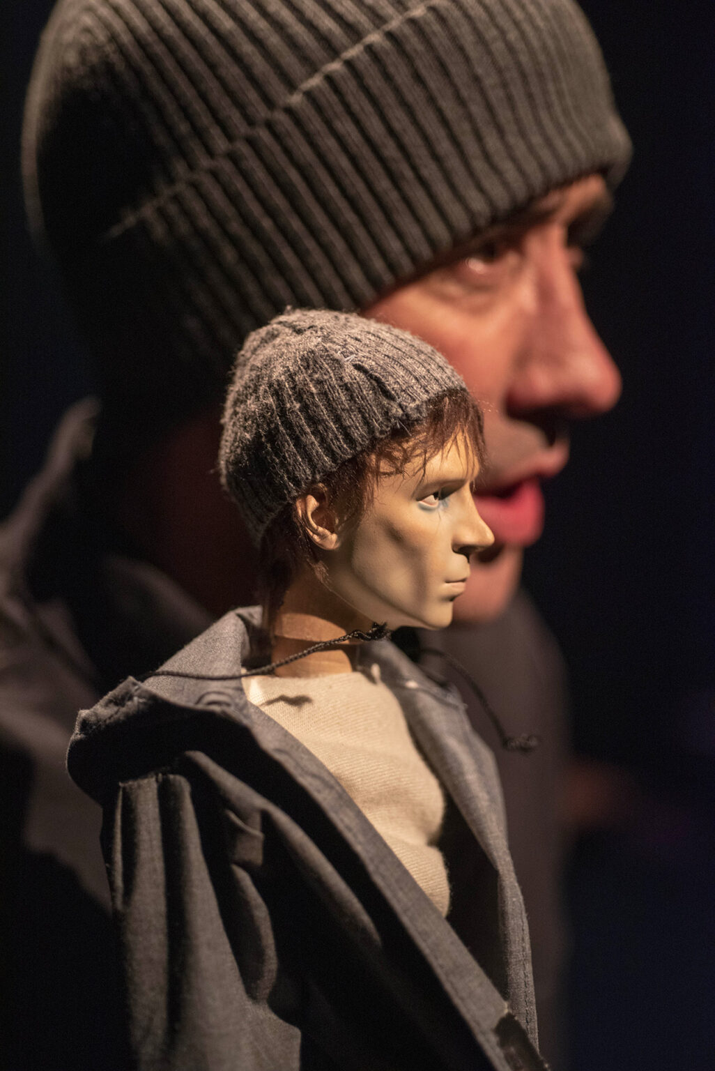 Une petite poupée d'homme portant un bonnet et une veste. Derrière lui, on voit le visage de l'acteur Cyril Briand qui lui aussi porte un bonnet.