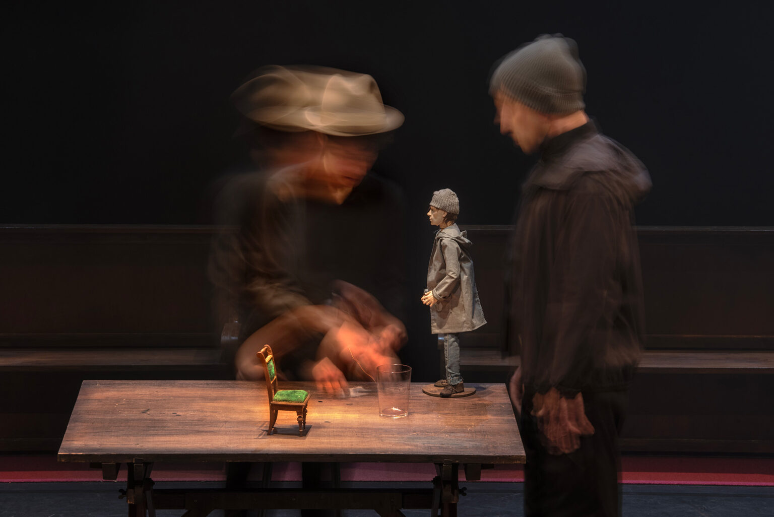 L'acteur Cyril Briand et l'actrice Marie Delhaye se font face de part et d'autre d'une table. Ils sont habillés de noir et porte bonnet pour l'un et chapeau pour l'autre. Sur la table, il y a une petite chaise, un verre et la figurine de Raskolnikov. Les acteurs, en plein mouvement, sont flous. Seuls la chiase et Raskolnikov se démarquent.