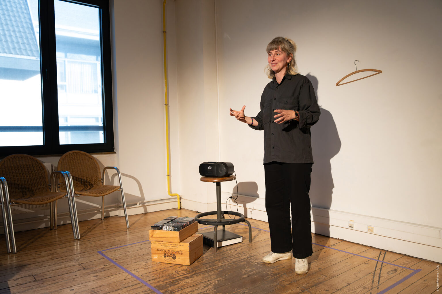 Cécile Hupin est debout. Elle porte une chemise et un pantalon noirs. A côté d'elle, un bac avec des cassettes audio et une radio.