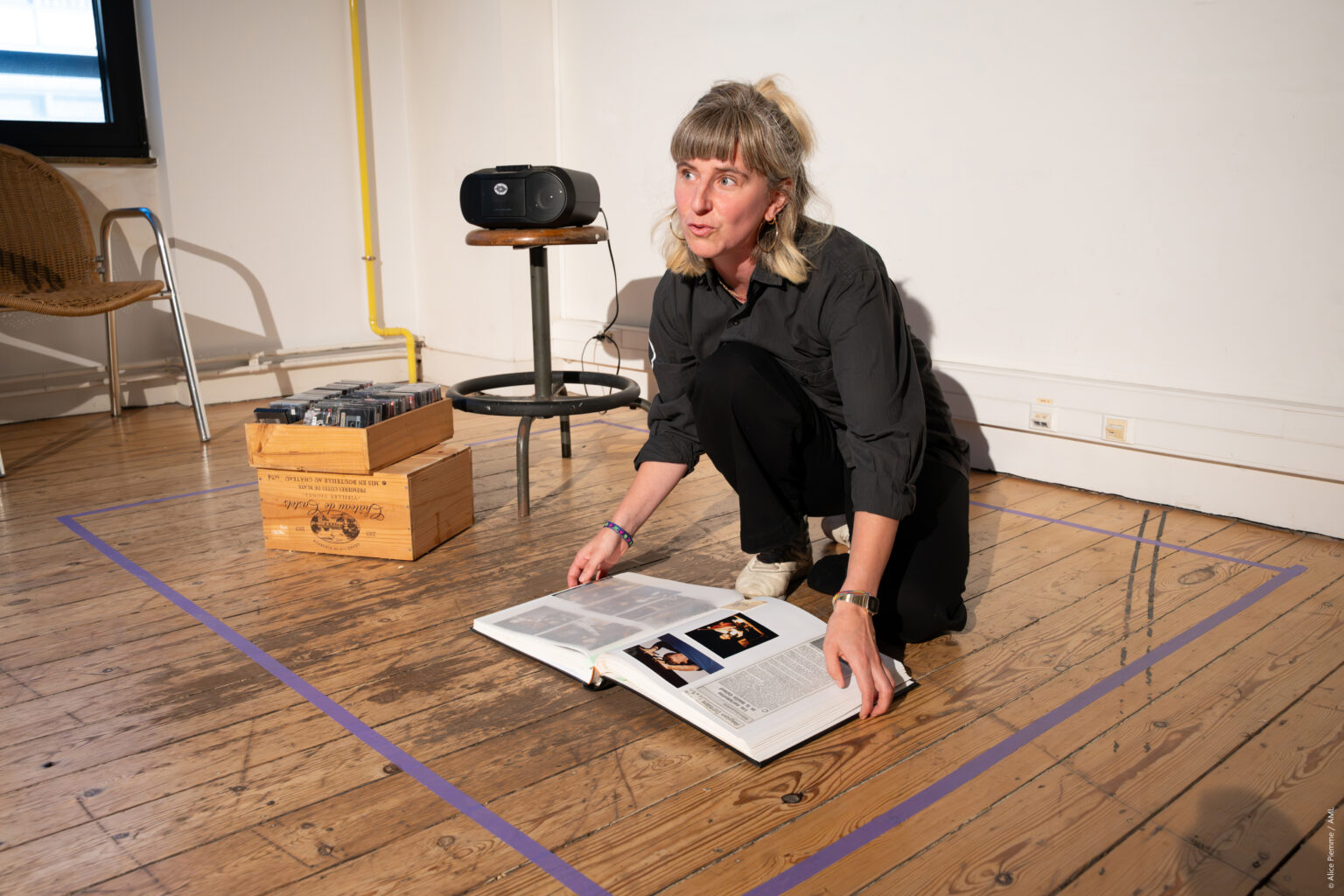 Cécile Hupin est assise devant un album reprenant des extraits de journaux. Elle porte une chemise et un pantalon noirs. A côté d'elle, un bac avec des cassettes audio et une radio.