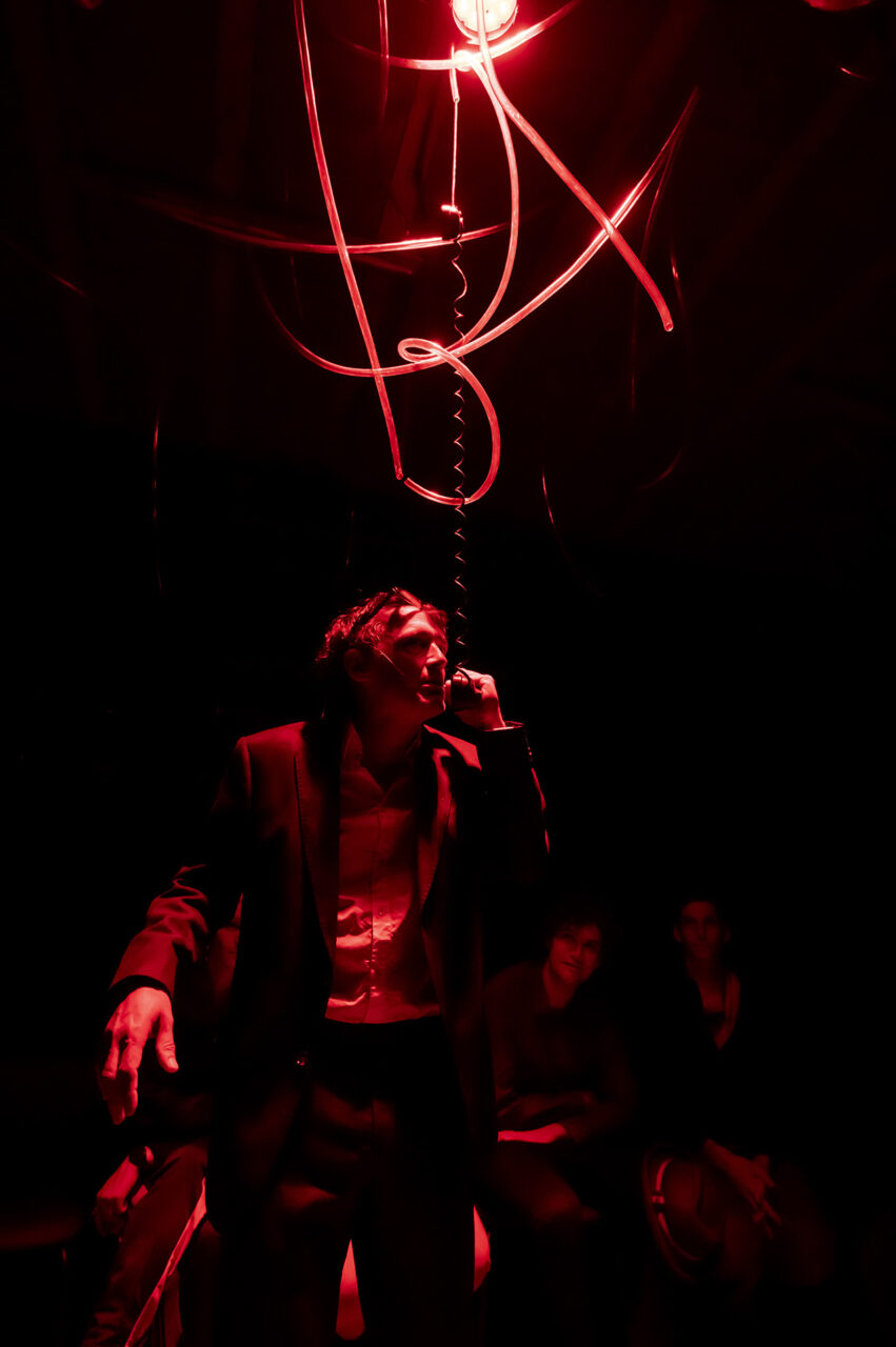 L'acteur Vincent Minne est debout, dans la semi-obscurité. Il n'est éclairé que par une lumière rouge. Il porte un costume et tient à la main un téléphone (ancien) qui est suspendu (accroché par un fil au-dessus).