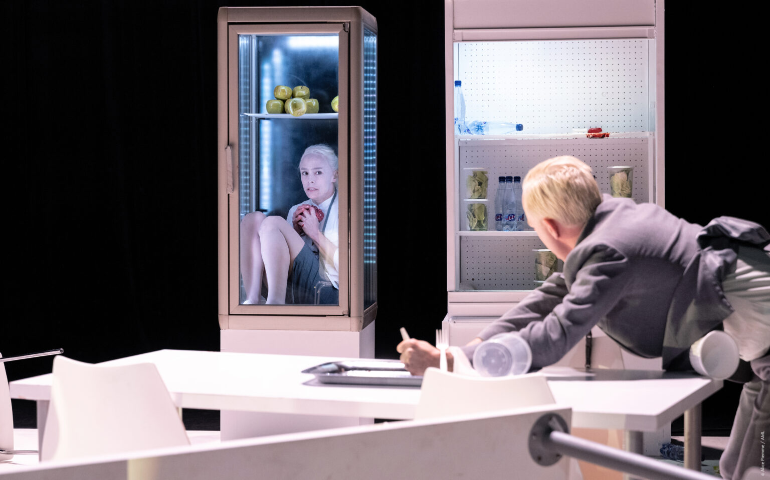 L'actrice Sophie Leso est blottie dans un petit frigo transparent. Elle tient un coeur dans ses mains. Aurelio Mergola la regarde et n'a pas l'air contente. Ils sont tous les deux habillés comme des employés de bureaux et ont les cheveux très blonds, presque blancs. Ils sont dans un décor de cantine.