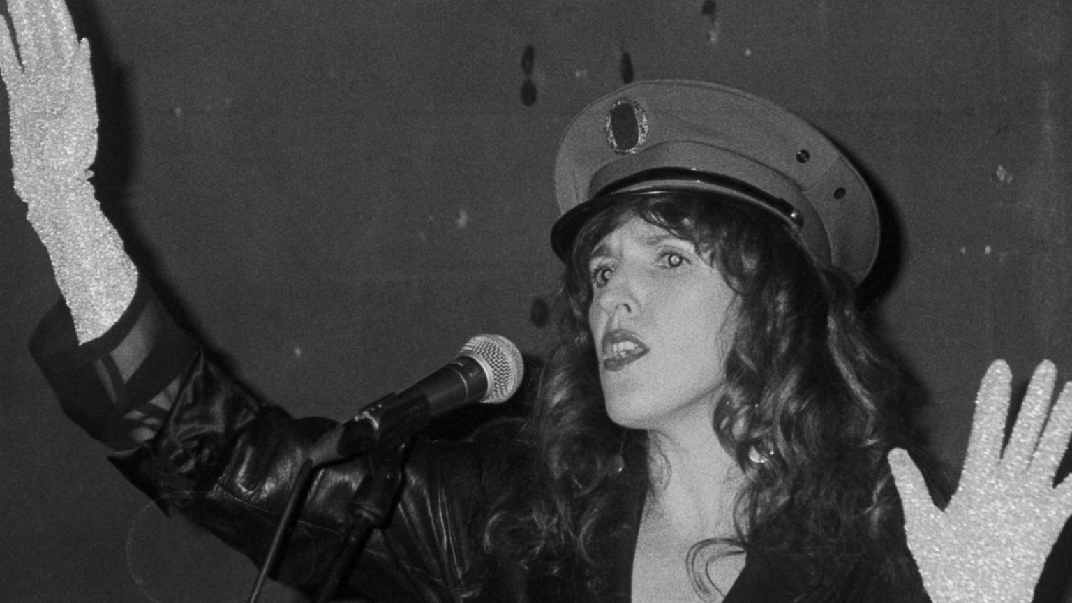 Photo en noir et blanc de Sarah Espour devant un micro. Elle porte une casquette type militaire et un gant argenté.