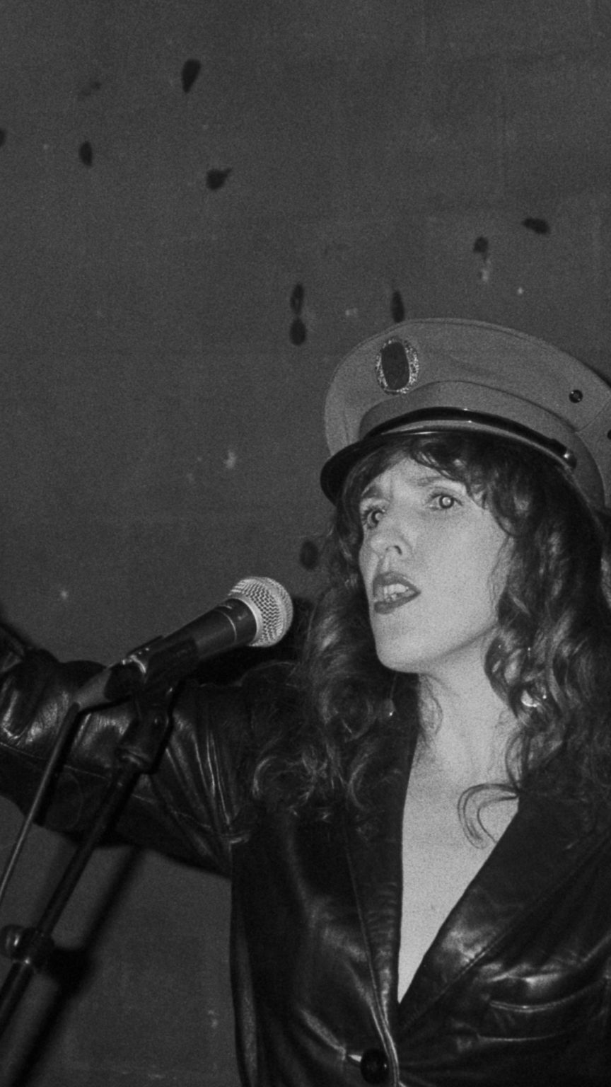 Photo en noir et blanc de Sarah Espour devant un micro. Elle porte une casquette type militaire et un gant argenté.