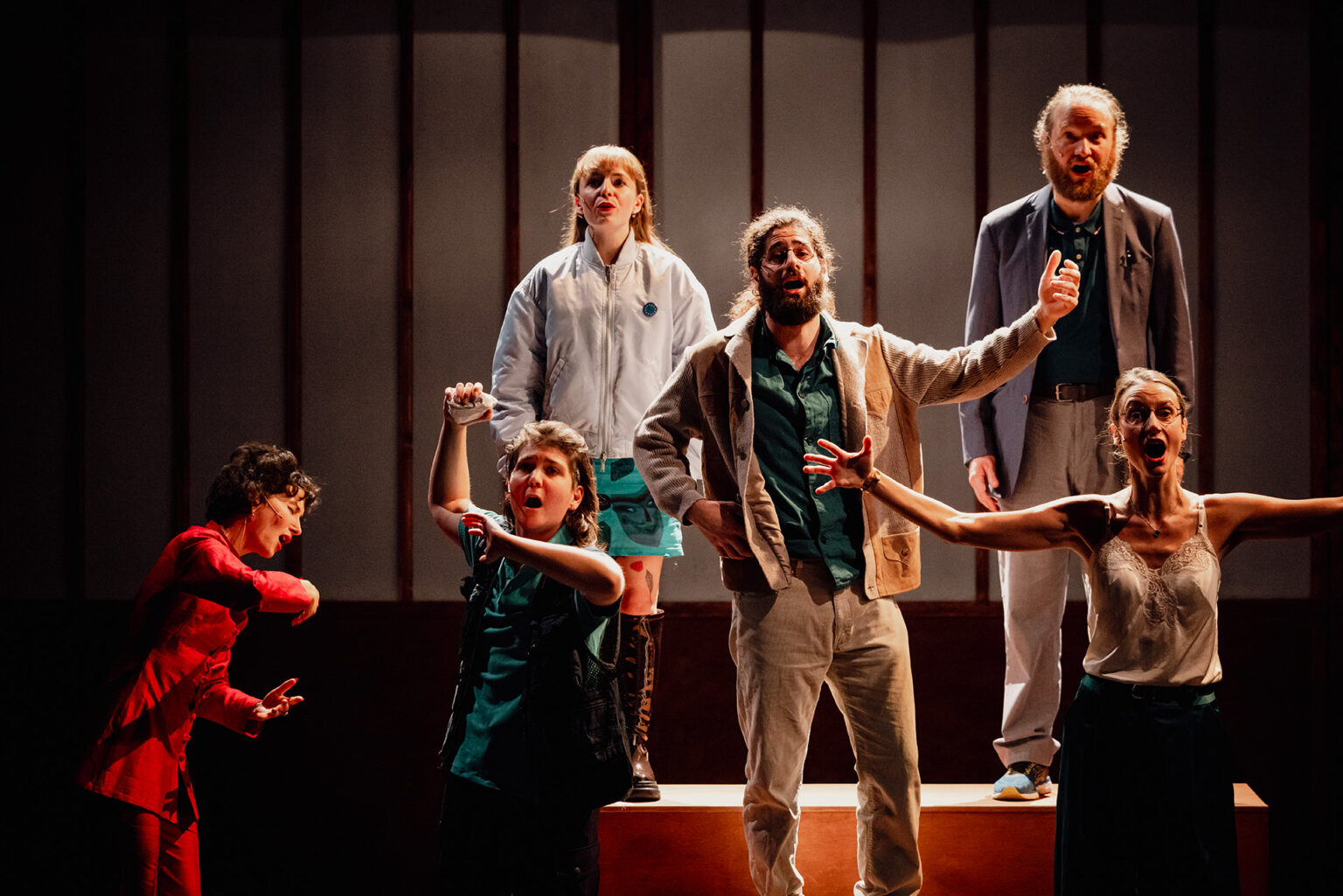 Sur une petite estrade, la chorale composée de cinq chanteurs répète. Ils prennent tous des poses étranges, type statues. Derrière eux, il y a un grand paravent en bois. Sur la gauche, la cheffe de choeur chante avec eux, à fond dedans.