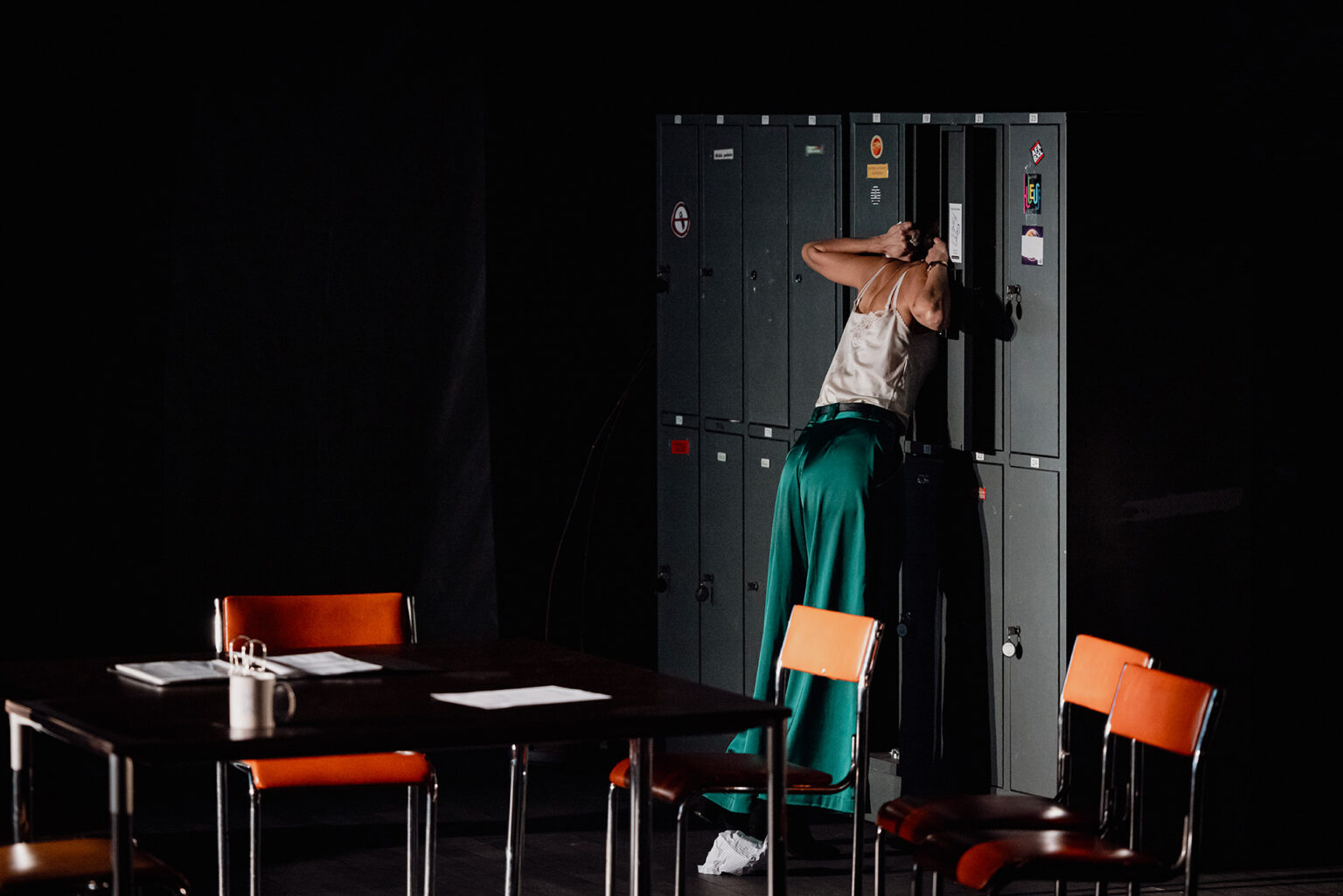 Une actrice qui porte un pantalon vert et un haut blanc met la tête dans un casier gris (type casier d'école). Autour d'elle, quelques chaises et tables.