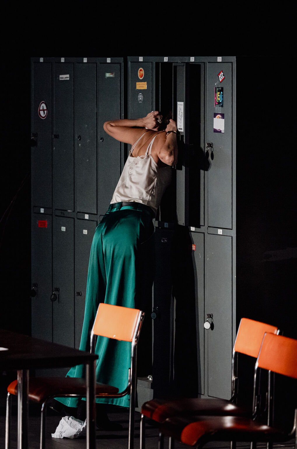 Une actrice qui porte un pantalon vert et un haut blanc met la tête dans un casier gris (type casier d'école). Autour d'elle, quelques chaises et tables.