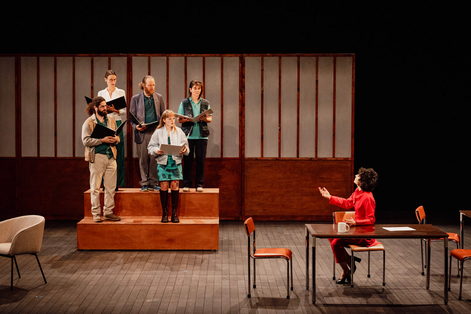 Sur une petite estrade, la chorale composée de cinq chanteurs répète, leurs partitions en mains. Derrière eux, il y a un grand paravent en bois. Sur la droite, la cheffe de choeur semble plus intéresser par ses chaussures que par eux.