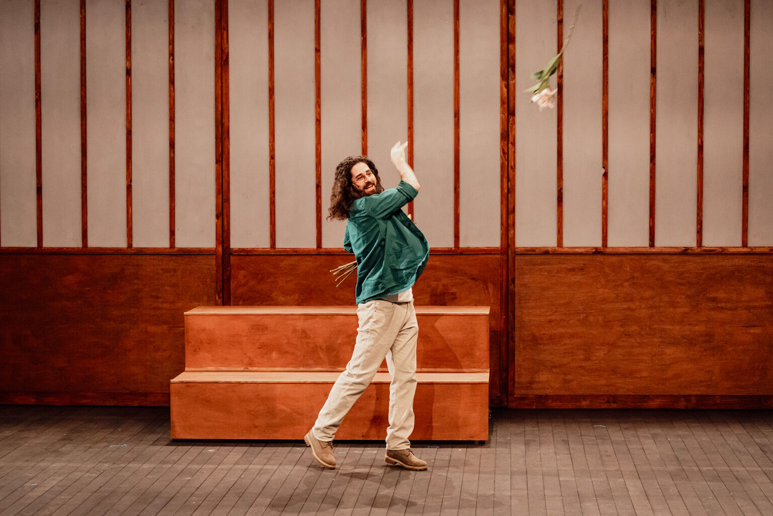Un acteur lance une fleur en l'air. Il porte une veste verte. Dernière lui, il y a un grand paravent en bois.