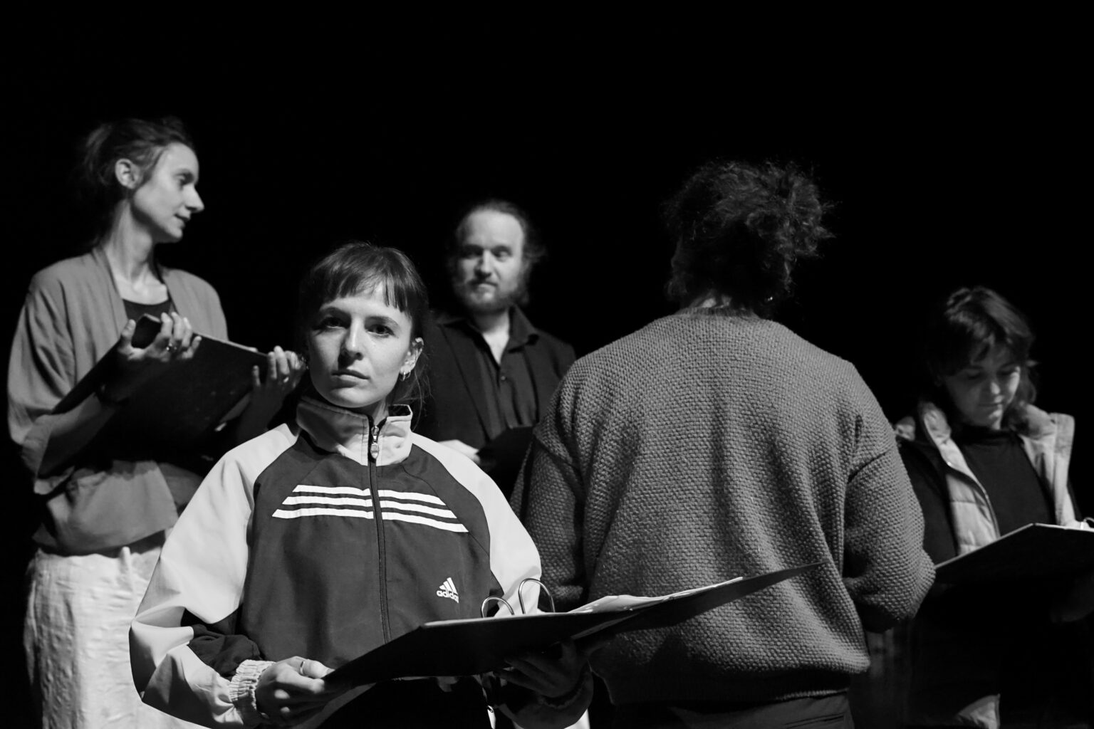 Photo en noir et blanc où l'on voit les participants de la chorale en préparation. L'une regarde la caméra, sa partition en mains.