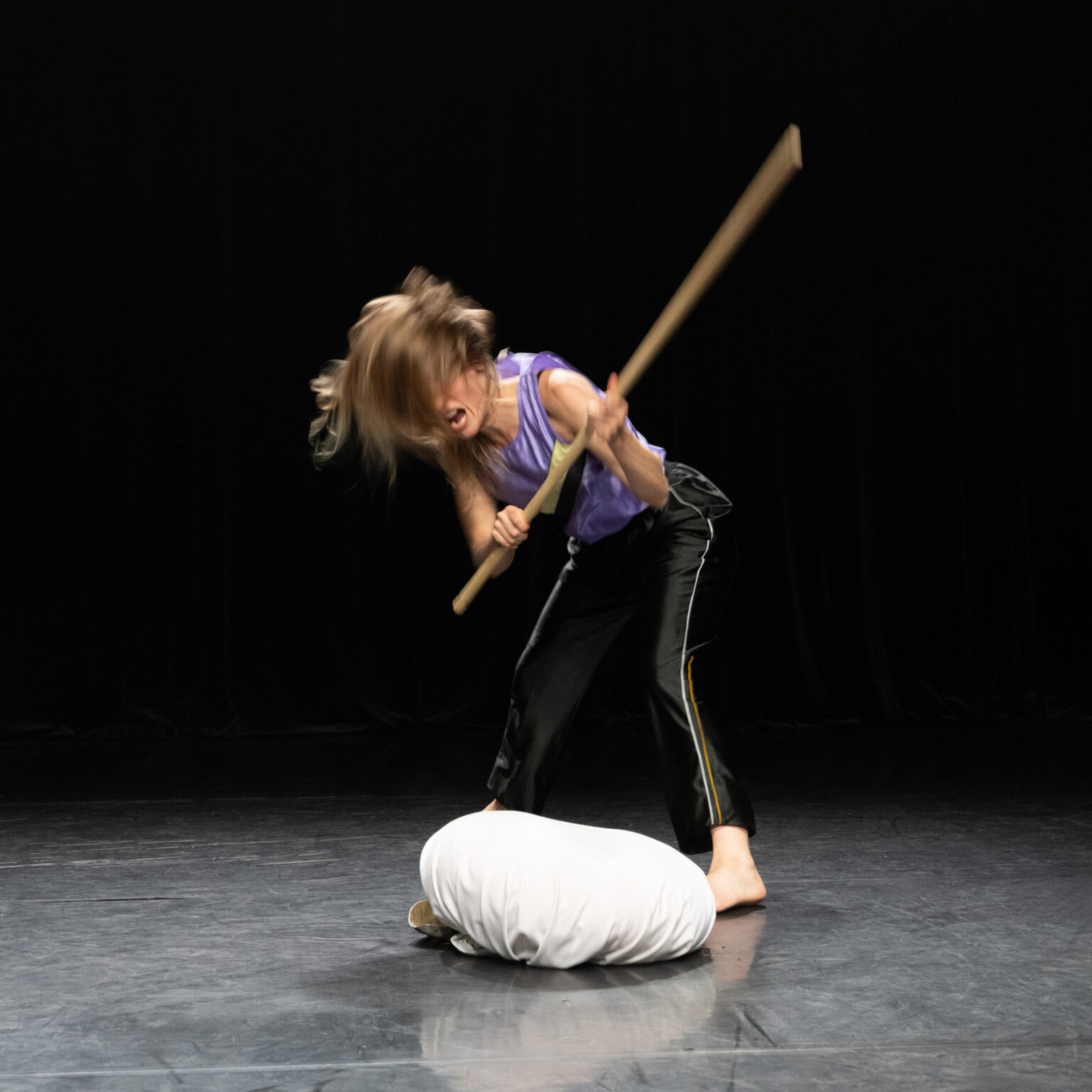 L'actrice Lucie Debay, habillée d'un jogging et d'une blouse à manches courtes mauve, tient un baton en mains et tape fort sur un coussin. Ses cheveux volent au vent.