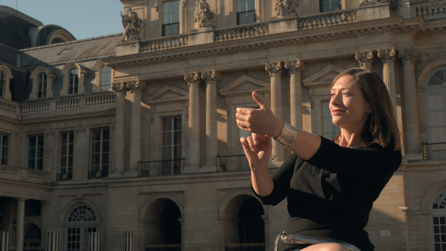 Ariane Loze pose en extérieur, devant un bâtiment style Renaissance. Elle porte une robe noire et touche sa main gauche avec 2 doigts de sa main droite.