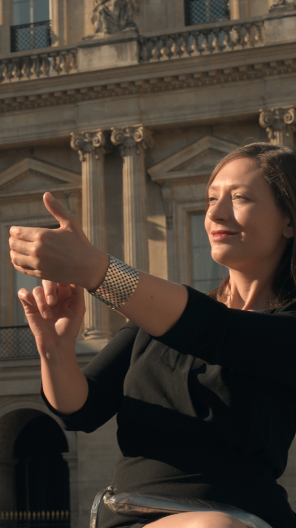 Ariane Loze pose en extérieur, devant un bâtiment style Renaissance. Elle porte une robe noire et touche sa main gauche avec 2 doigts de sa main droite.