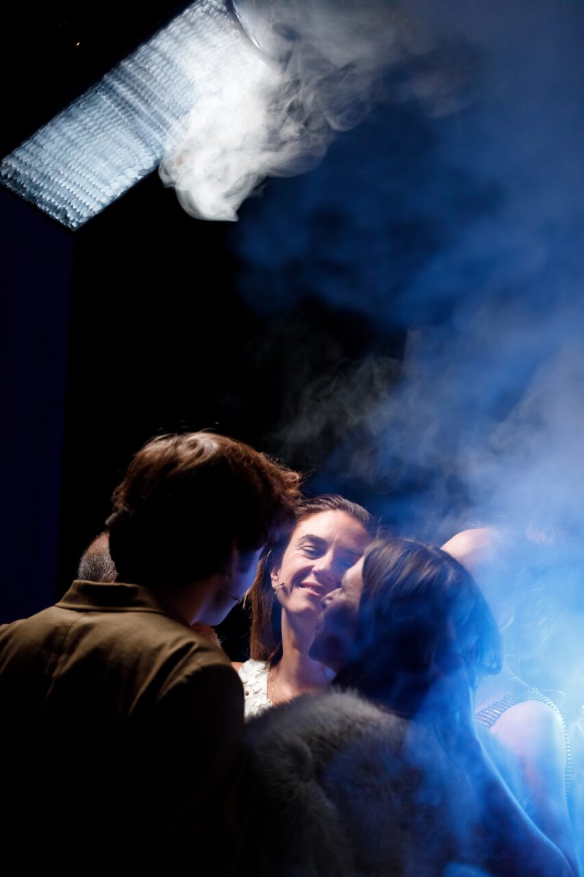 Dans une semi-obscurité, quelques acteurs et actrices se font face. On voit le visage de l'une des actrices, éclairée par un projecteur, elle sourit.