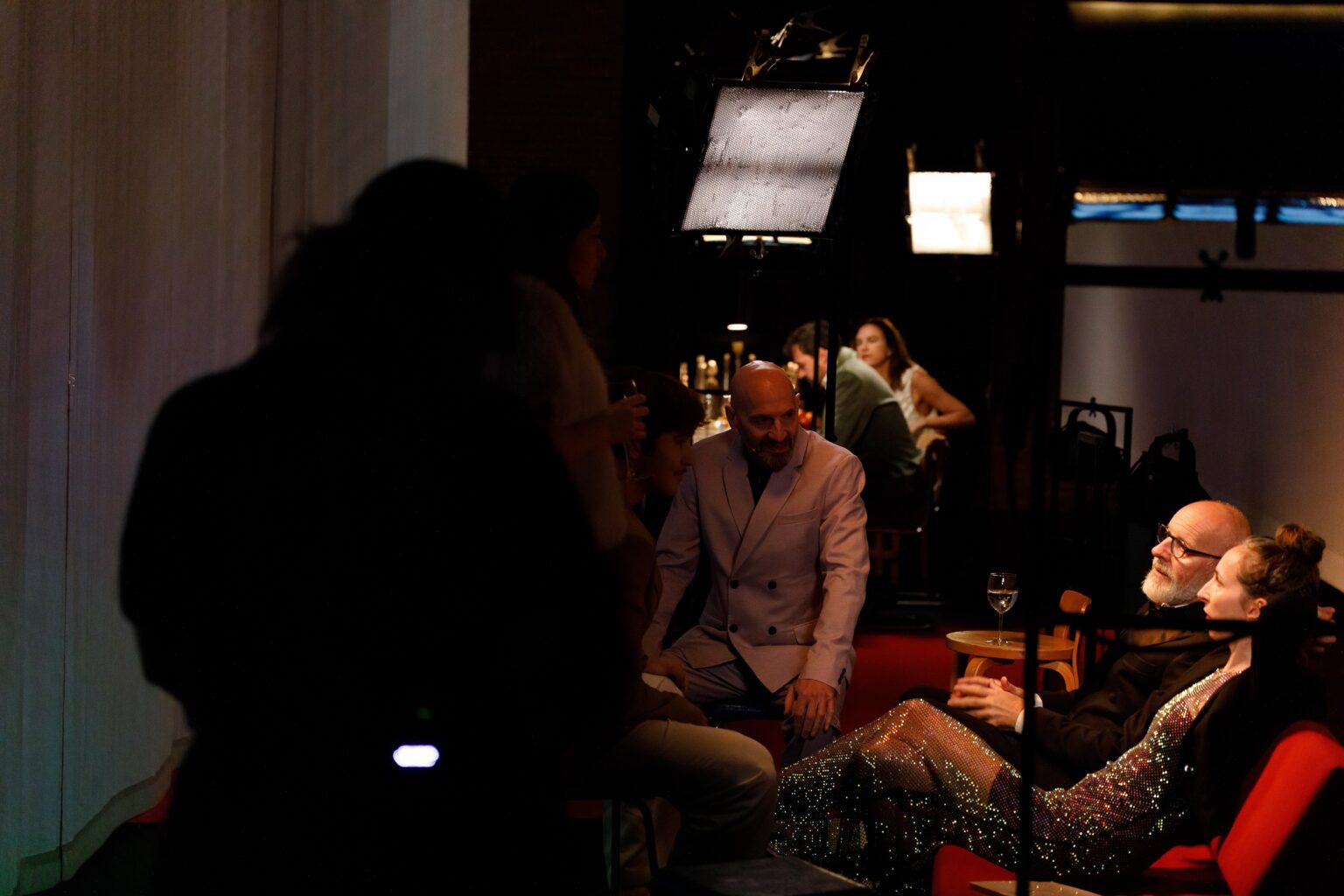 Les acteurs et actrices de Opening Night sont installés dans un canapé rouge ou autour d'une table à l'arrière. Ils sont filmés et éclairés par des projecteurs de cinéma.