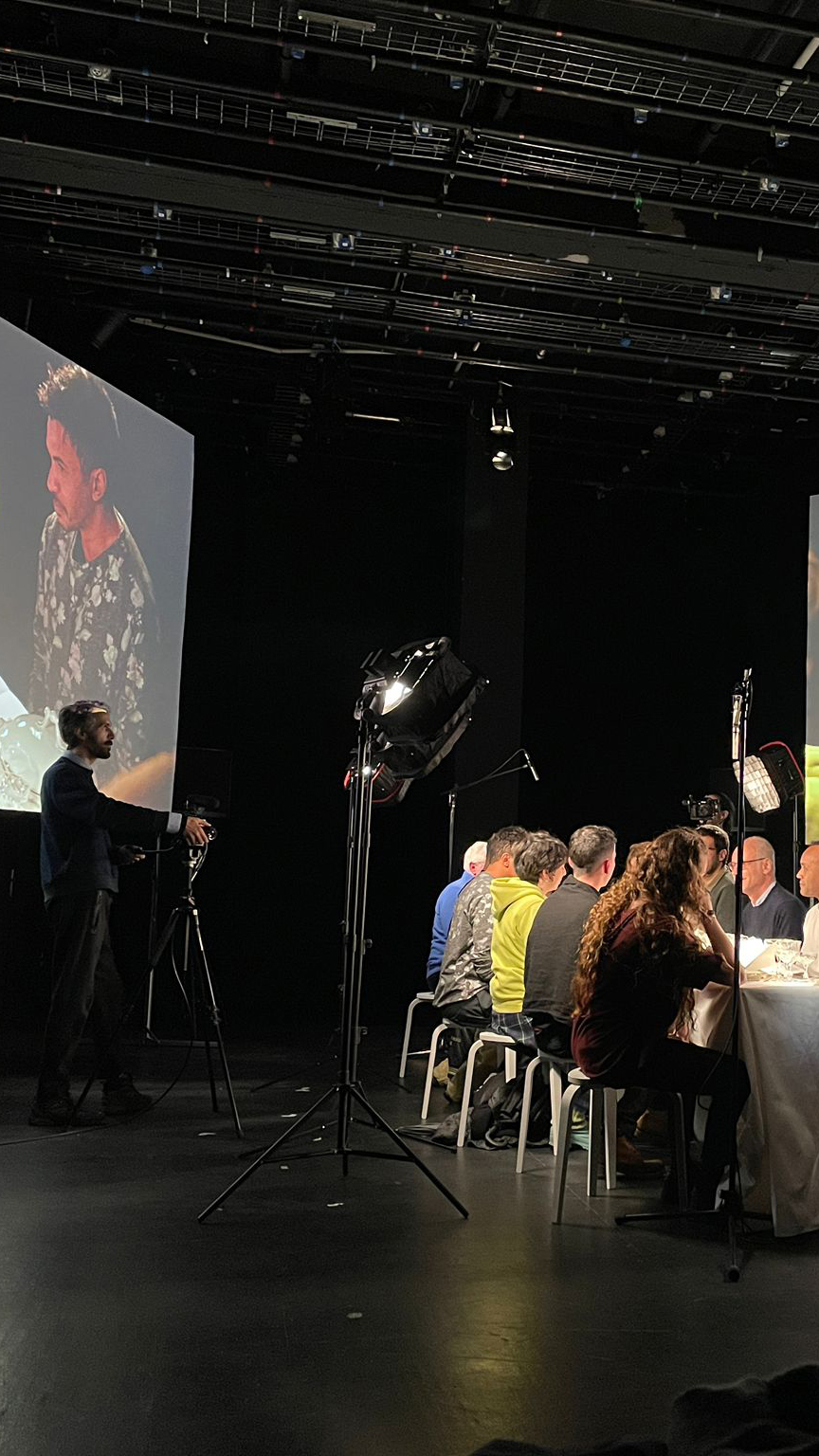 Sur le plateau, il y a une grande table où sont attablés des personnes du public. Deux grands écrans encadrent le plateau.
