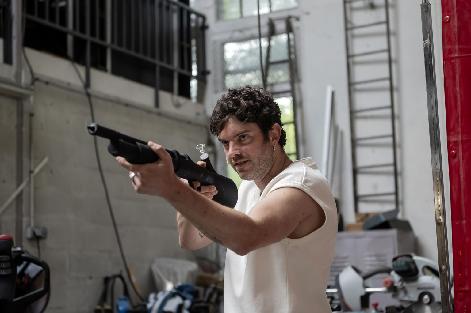Un acteur tient une arme en mains et vise quelqu'un. Il est dans ce qui semble être un atelier. Il porte un t-shirt blanc.