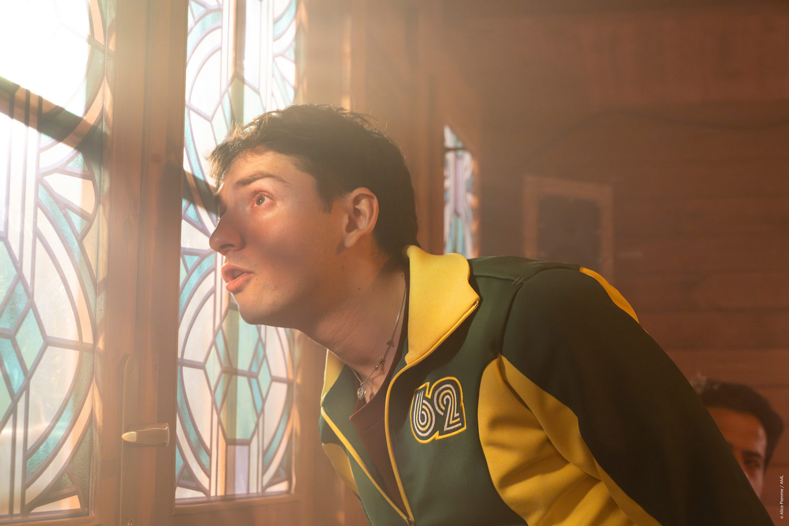 Un jeune comédien regarde par la fenêtre d'un chalet en bois. Il porte une veste de survêtement jaune et vert.