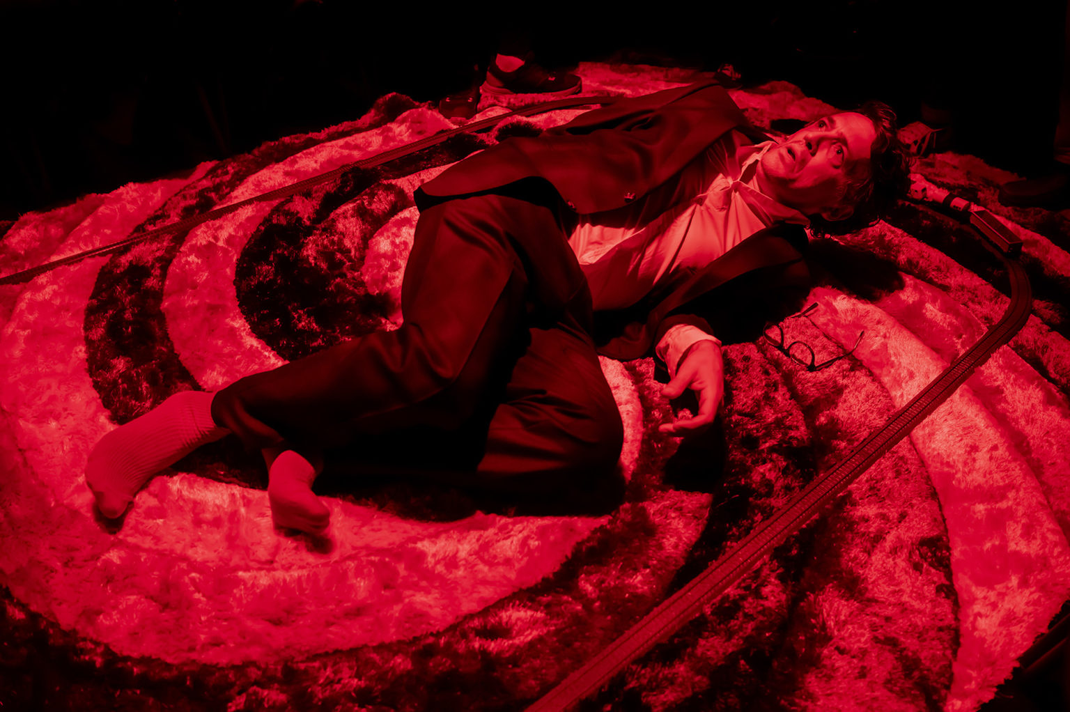 Un homme est au sol, sur un tapis, entouré d'un train électrique. Il a l'air apeuré. Il porte un costume. Il est éclairé par une lumière rouge.