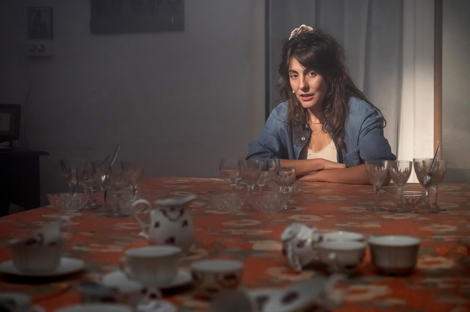 Une actrice est installée autour d'une table sur laquelle sont disposées un service à thé et des verres. Il y a une nappe style années 60 avec des fleurs.