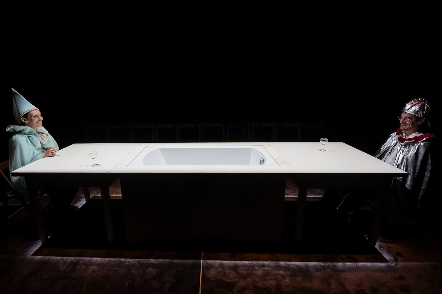 Un acteur (habillé en chevalier) et une atrice (habillée en fée) se font face autour d'une longue table au milieu de laquelle il y a une sorte de baignoire