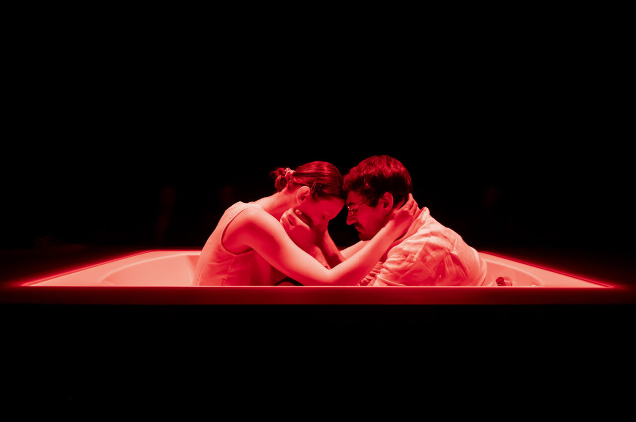 Un acteur et une actrice sont assis dans une baignoire face à face. Leurs visages se frôlent et reposent l'un sur l'autre, tout comme leurs mains posées sur la nuque de l'autre. Ils sont éclairés par une lumière rouge.