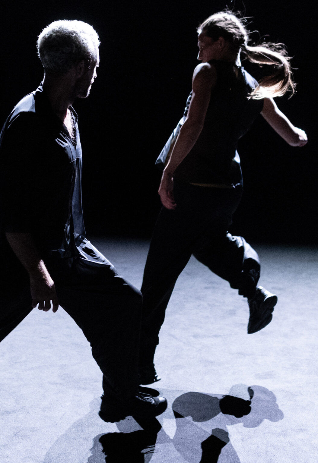 Angela Rabaglio et Micaël Florentz dansent l'un à côté de l'autre, en habits foncés. Le tapis de danse est clair. Il a l'air de marcher autour d'elle pendant qu'elle sautille, et lève l'un de ses pieds.