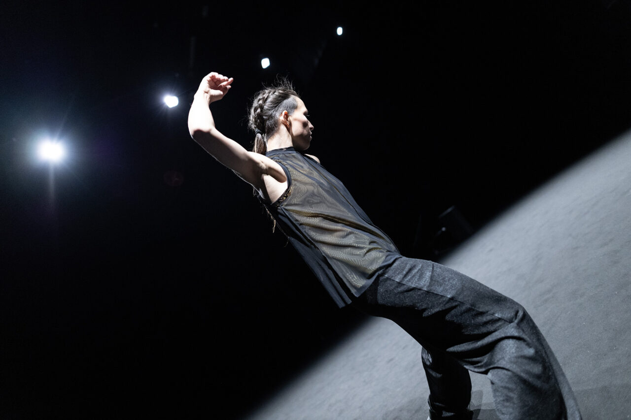 Angela Rabaglio est de dos, le bras droit relevé au-dessus de sa tête. Elle porte des vêtements sombres.