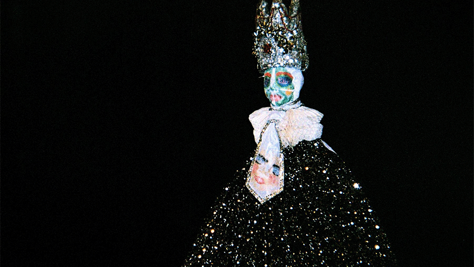 Devant un fond noir se tient une figure dans une longue robe noire étincelante, elle porte un masque bleuté et un chapeau pointu brillant