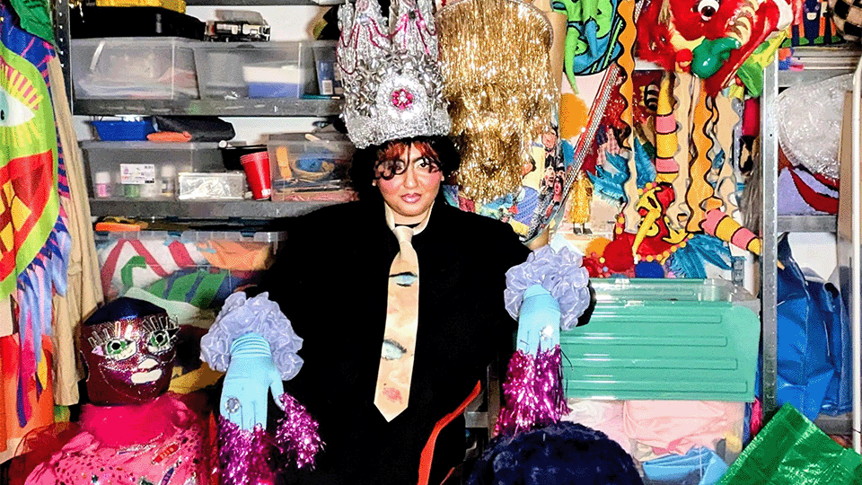 Une personne est assise au milieu d'une pièce très colorée, elle porte un chapeu et des gants au design très coloré et contemporain. Elle est entourée de masques et autres objets artisitques.