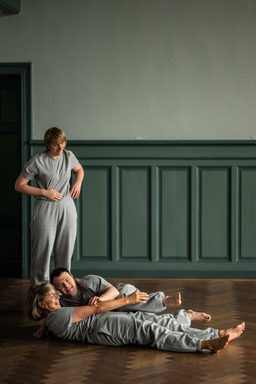 Deux danseurs sont couchés au sol, dans les bras l'un de l'autre et ils rient. Une autre est debout proche d'eux et sourit aussi. Pris en photo comme si c'était un moment de rigolade. Ils sont dans une salle au parquet en bois et aux murs peints en vert. Tous sont habillés en gris.
