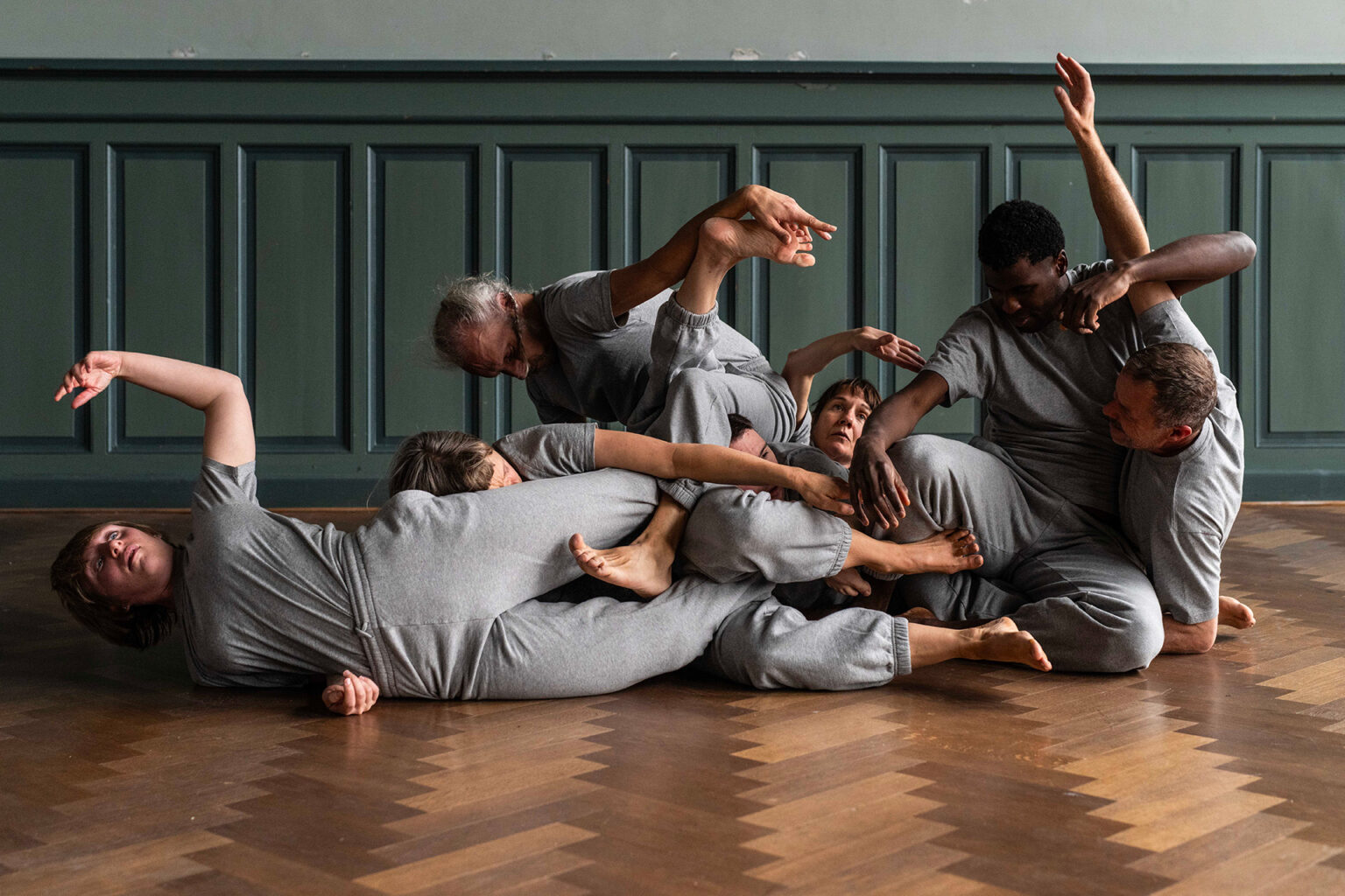 Les danseurs, tous habillés de gris, sont couchés ou en position assises sur le sol. Leurs corps s'imbriquent les uns aux autres.