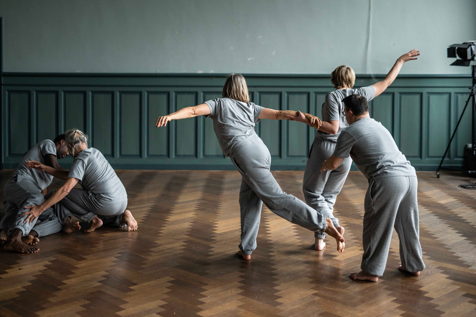Côté gauche, deux danseurs sont assis, imbriqués l'un à l'autre. De l'autre côté, trois danseurs sont en position debout. Ils se tiennent la main  et font quelques pas de danse. Ils sont dans une salle au parquet en bois et aux murs peints en vert.