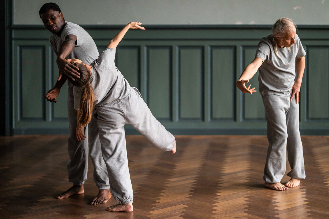 Un danseur et une danseuse sont debout et tous les deux habillés avec un jogging et un t-shirt gris. La danseuse lève un bras et une jambe. L'autre la retient avec sa main par la tête. Ils sont dans une salle au parquet en bois et aux murs peints en vert.