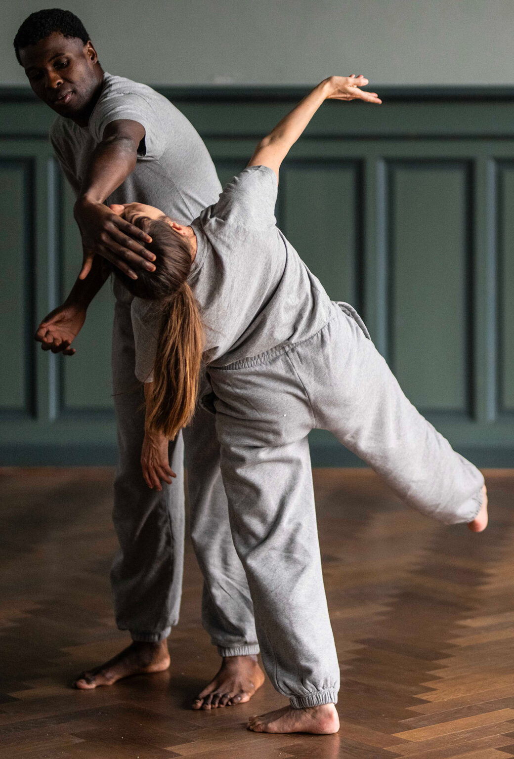 Un danseur et une danseuse sont debout et tous les deux habillés avec un jogging et un t-shirt gris. La danseuse lève un bras et une jambe. L'autre la retient avec sa main par la tête. Ils sont dans une salle au parquet en bois et aux murs peints en vert.