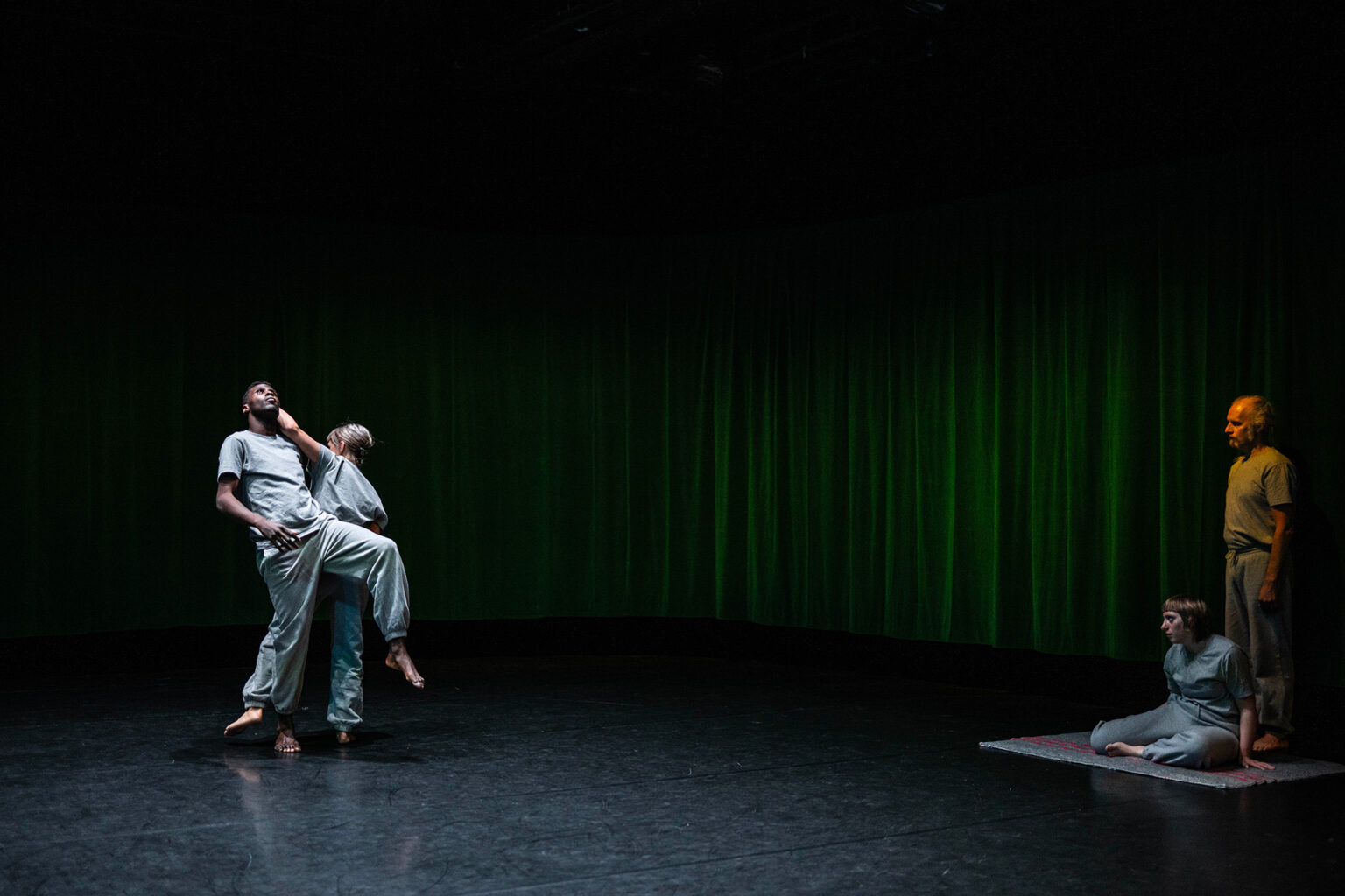Deux danseurs dansent en duo à gauche, collés l'un contre l'autre. Deux autres les regardent. ls portent des vêtements gris (pantalon de training et t-shirt).