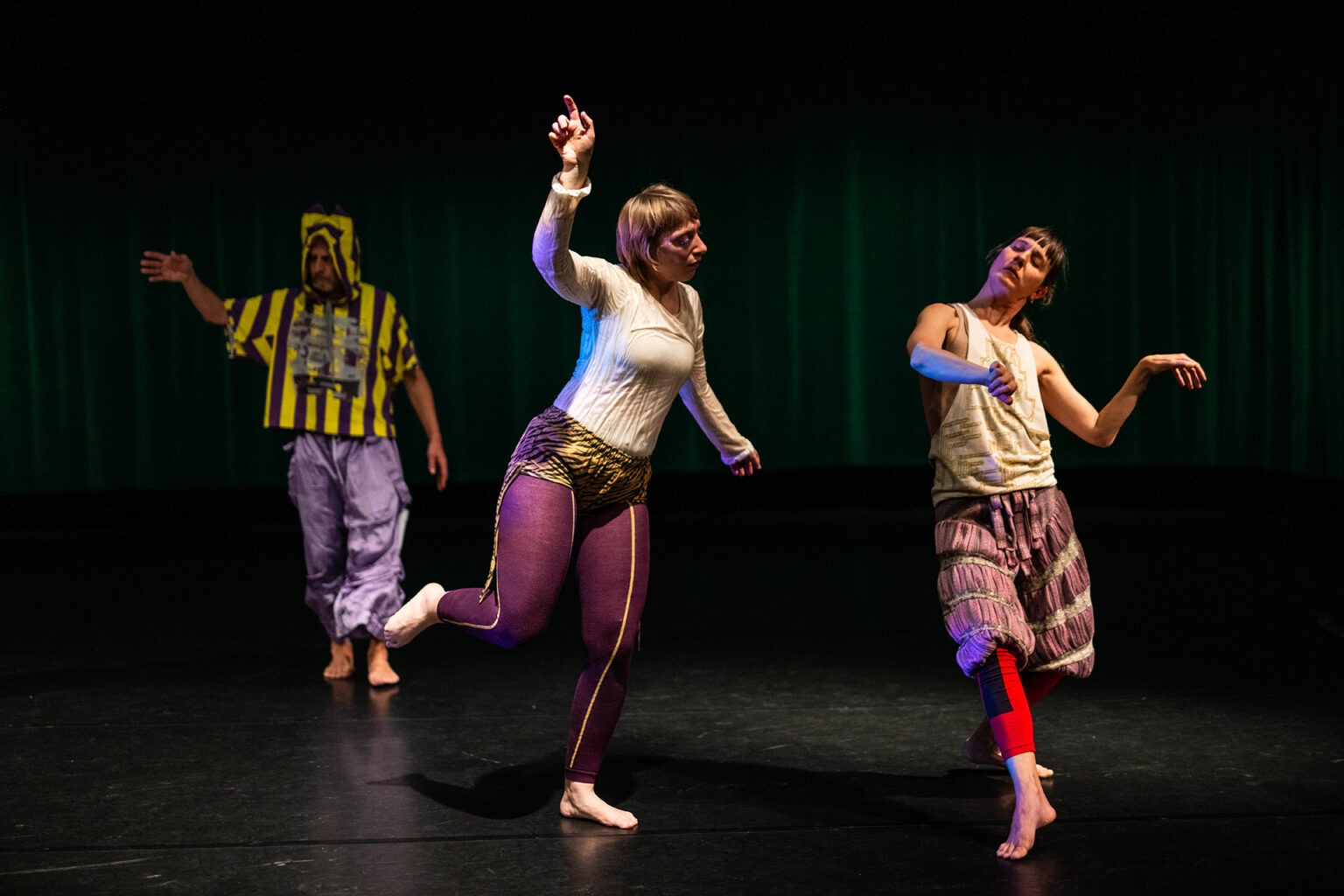 Trois danseurs dansent, les mains en l'air. Ils portent des vêtements bariolés.
