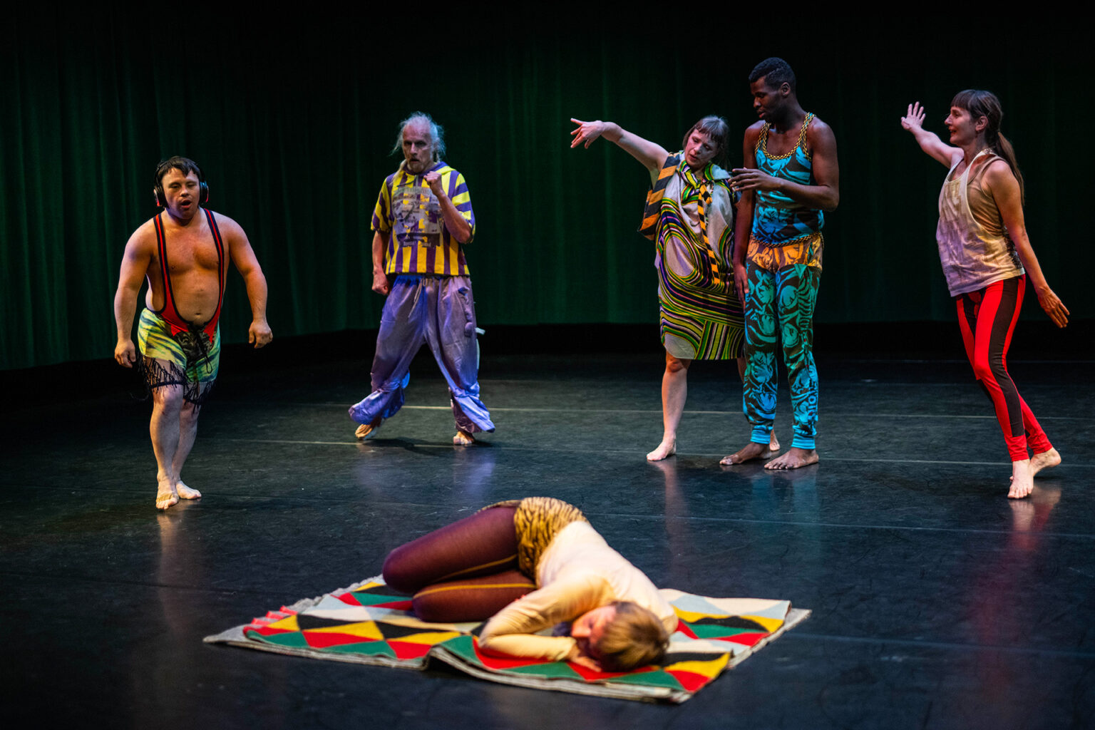 5 danseurs sont debout, à l'arrière scène. Ils portent des vêtements bariolés. À l'avant-scène, l'une des danseuses est couchée au sol, sur une couverture bariolée.